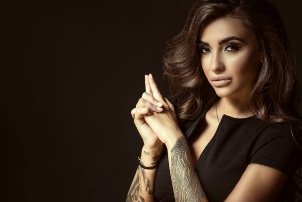 Portrait of young beautiful tattooed woman with luxuriant shining wavy hair and perfect make up holding her hands in shooting gesture in the shape of the gun - Foto, Imagem