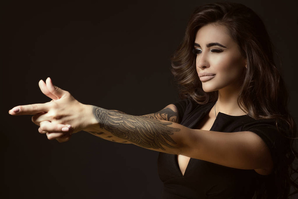 Portrait of young beautiful tattooed woman with luxuriant shining wavy hair and perfect make up pretending to aim something with shooting gesture of her hands - Photo, Image