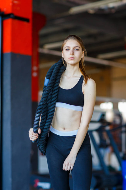 An attractive young woman standing in a fitness center posing with a physical rope over her shoulder. - Φωτογραφία, εικόνα