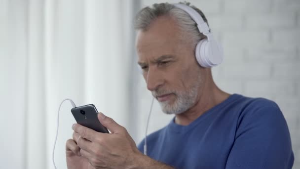 Senior man in headphones scrolling screen of smartphone, easy to use application - Πλάνα, βίντεο