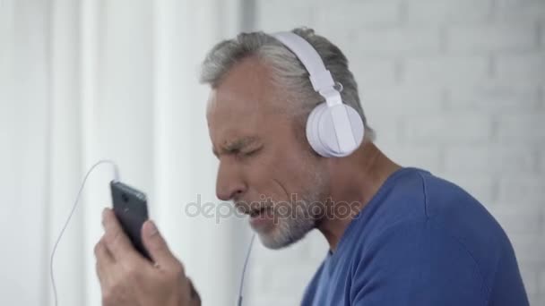 Male retiree in headphones enjoying favorite song from cellphone and dancing - Felvétel, videó