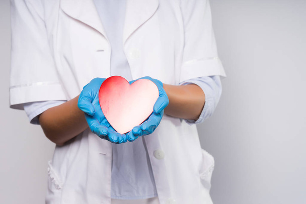 a girl in medical clothes and gloves holds a paper heart in her hands. The heart is colored by a gradient from red to white - Fotografie, Obrázek