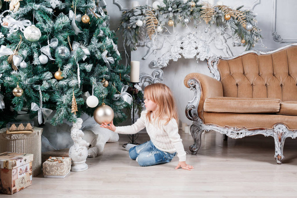 Adorable niña rubia en un vaquero azul sentada en un piso cerca del árbol de Navidad y mirando un juguete
. - Foto, imagen