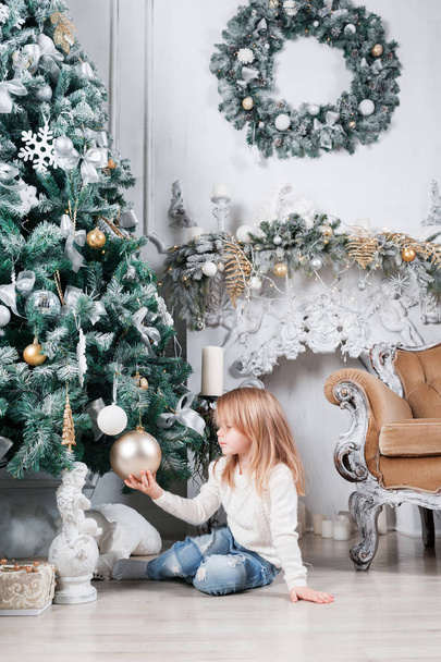 Adorable little blonde girl in a blue jeans sitting on a floor near christmas tree and looking on toy. - Valokuva, kuva