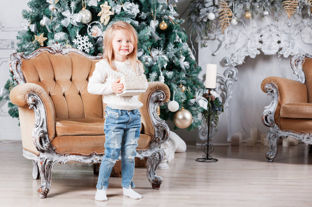 χαριτωμένο μικρό κορίτσι που κάθεται σε μια καρέκλα και ανοίγει ένα κουτί με ένα δώρο για φόντο χριστουγεννιάτικο δέντρο με στολίδια. - Φωτογραφία, εικόνα