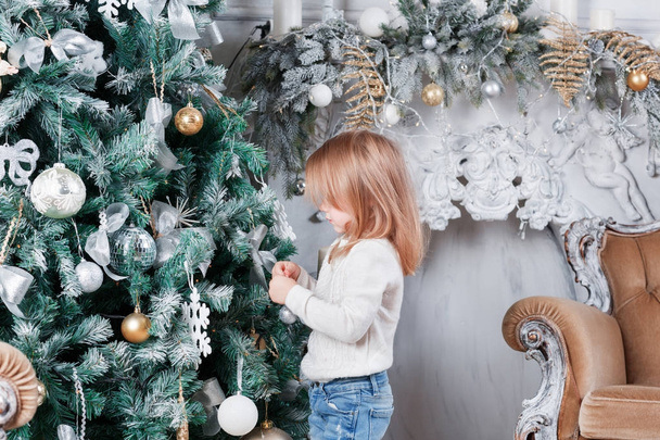 Αξιολάτρευτο μικρό ξανθό κορίτσι σε ένα μπλε τζιν κάθεται στο πάτωμα κοντά στο χριστουγεννιάτικο δέντρο και να ψάχνει για παιχνίδι. - Φωτογραφία, εικόνα