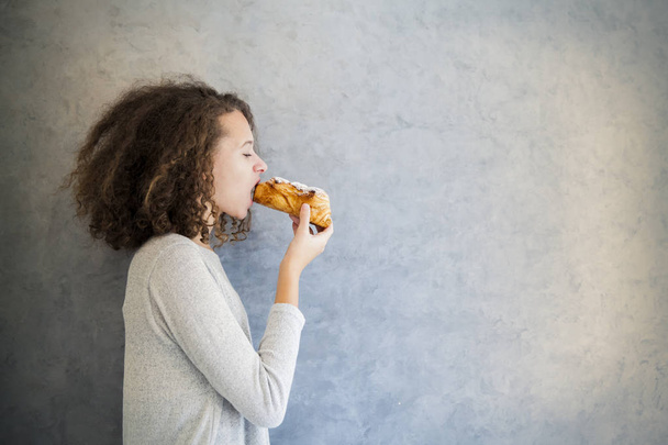 Cura cabelo encaracolado menina comer croissant contra a parede
 - Foto, Imagem