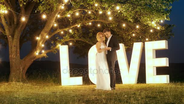 Молодая пара влюбленных в вечерние платья танцуют возле любовных светлых букв
. - Кадры, видео