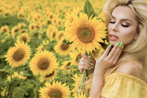 Kaunis blondi nainen seisoo niityllä auringonkukkien kanssa ja hymyilee onnellisesti
 - Valokuva, kuva