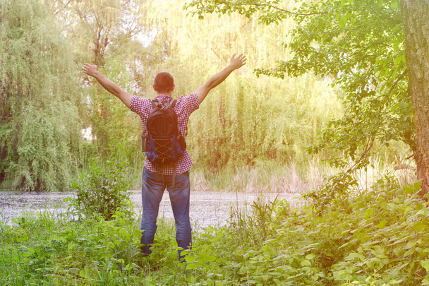 Парень с рюкзаком стоит на берегу лесного озера с распростертыми руками, с видом сзади
 - Фото, изображение