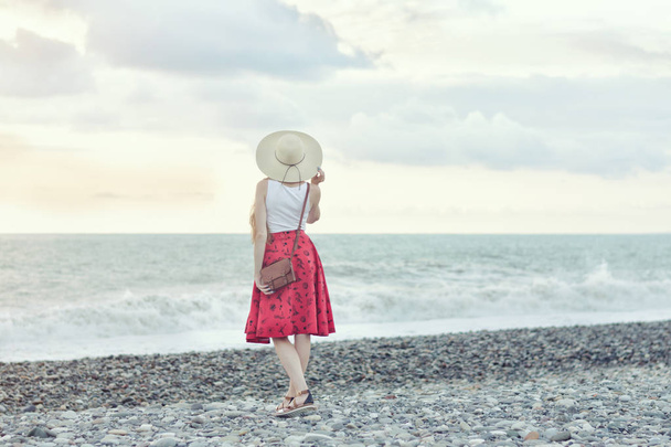 Tyttö punaisessa hameessa ja hattu päässä seisoo meren rannalla, auringonlaskussa. Näkymä takaa
 - Valokuva, kuva