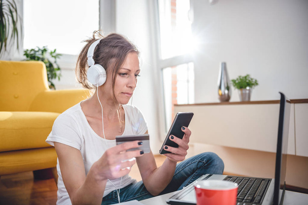 Woman wearing white shirt and headphones shopping online on smart phone and using credit card - Foto, Imagem