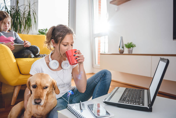 Woman wearing white shirt sitting on the floor by the table drinking coffee and petting a dog - 写真・画像