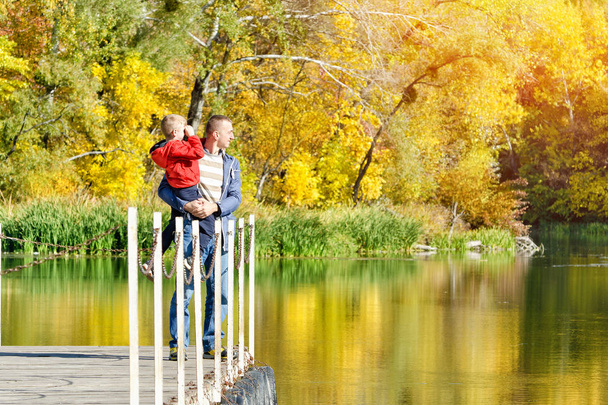 Отец и сын на руках стоят на причале у реки. Солнечная осень
 - Фото, изображение