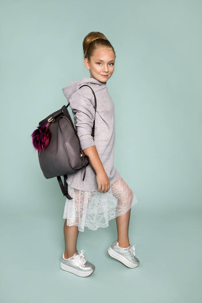 Ładna dziewczynka ubrana w szkole ubrania i szkolny plecak stojąc na niebieskim tle - Zdjęcie, obraz