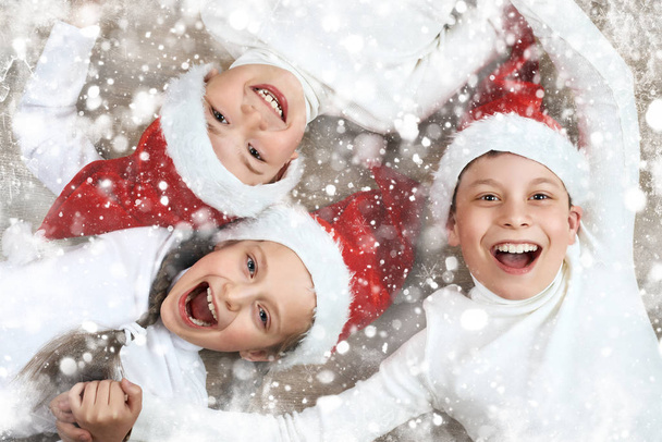 child group lying together on wooden background and laugh, dressed in christmas Santa hat and having fun, winter holiday concept, snow decoration - Photo, Image