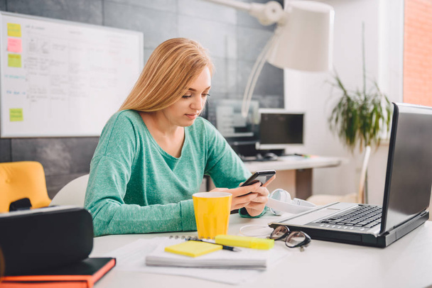 Женщина сидит за столом в офисе и пользуется смартфоном
 - Фото, изображение