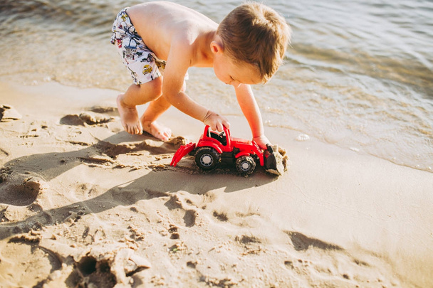 Junge spielt am Strand in der Nähe des Flusses mit rotem Traktor - Foto, Bild