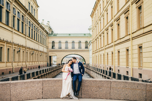 Porträt eines hübschen Bräutigams und einer Braut, die sich umarmen, auf einer alten Brücke sitzen, Ruhe und Zweisamkeit genießen, eine gute Beziehung haben. Hochzeit, Feier und Ehekonzept - Foto, Bild