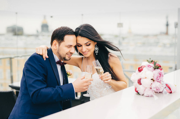 Young affectionate couple in love clink glasses with champagne, embrace each other as celebrate their wedding, being glad to become one family, enjoy togetherness, pose joyfully at camera - Photo, Image