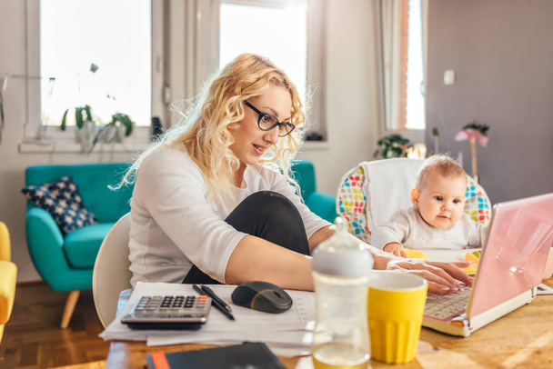 Mother wearing eyeglasses working at home office on laptop and taking care of her baby - Photo, image