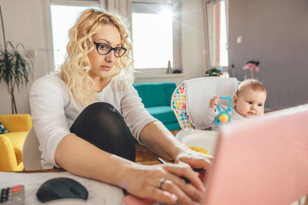 Mother wearing eyeglasses working at home office on laptop and taking care of her baby - Фото, изображение