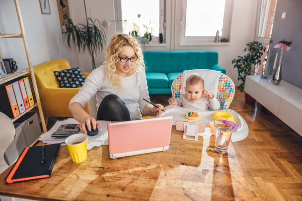 Mother wearing eyeglasses working at home office on laptop and taking care of her baby - Foto, Bild