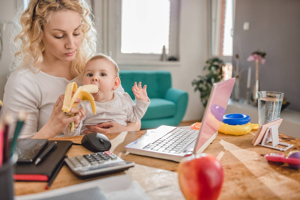 Mãe alimentando bebê em casa escritório com bananas
 - Foto, Imagem