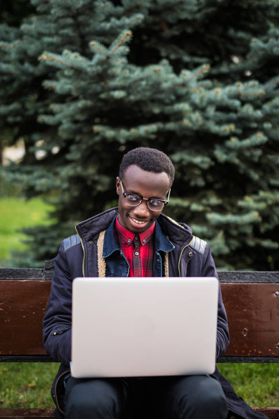 Foto di giovane uomo allegro africano seduto sulla panchina e utilizzando il computer portatile. Guardando la macchina fotografica
. - Foto, immagini