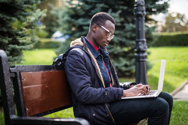 Joyful African american man using computer outdoors on the bench at street - Фото, изображение