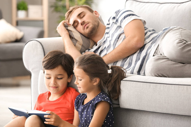 Cute children reading book while their father is sleeping on sofa at home - Foto, Bild