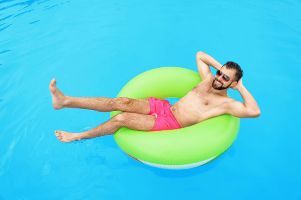 Handsome young man with inflatable ring relaxing in blue swimming pool - Photo, image