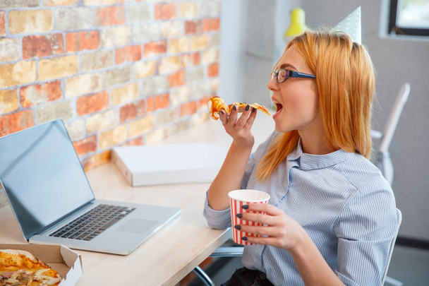 A girl eating pizza in the office in front of a laptop - 写真・画像