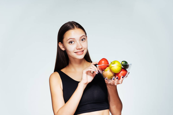 uma jovem bela menina de esportes observa sua figura, segura um prato com legumes de baixa caloria e frutas em suas mãos
 - Foto, Imagem