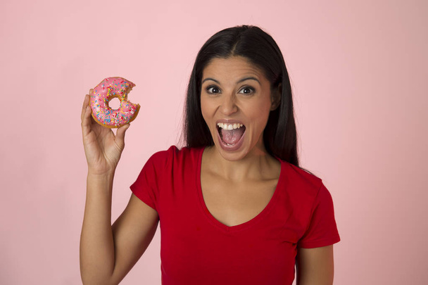 junge glückliche attraktive lateinische Frau in rotem Top lächelnd aufgeregt halten Zucker Donut isoliert auf rosa Hintergrund - Foto, Bild