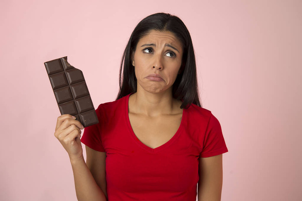 aantrekkelijk en gelukkig Spaanse vrouw in rode top schuldgevoel houden chocolade bar op roze achtergrond - Foto, afbeelding