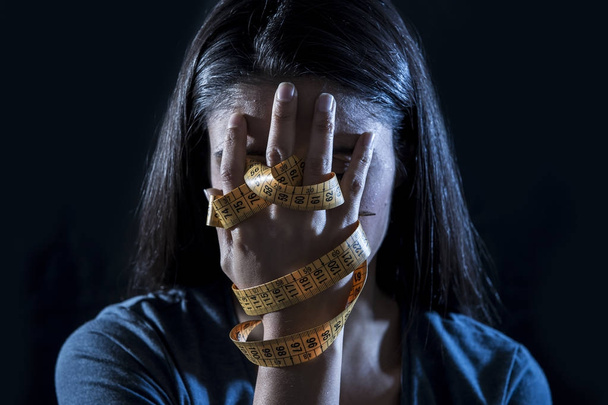 hands wrapped in tailor measure tape covering face of young depressed and worried girl suffering anorexia or bulimia nutrition disorder - Foto, afbeelding