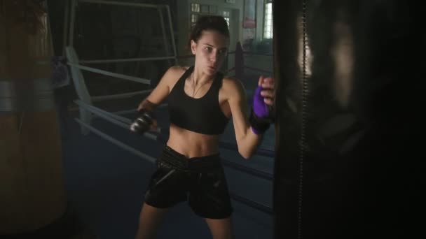 Athletic female boxer in gloves hiting boxing bag with her fist while training in a dark fitness studio with smoke. Slowmotion shot - Záběry, video