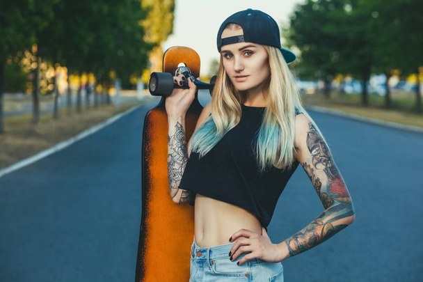 A beautiful, sexy hipster blonde with blue hair in tattoo stands with a longboard   - Photo, Image