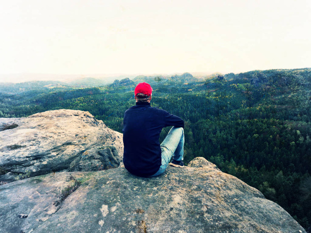 Természetjáró ember a szikla. Turista, fekete pulóver, farmer, piros sapkát ülni éles hegyi szikla-völgy felett - Fotó, kép