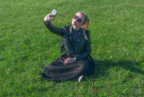 Beautiful blonde girl sitting on a green lawn and makes selfie - Fotoğraf, Görsel