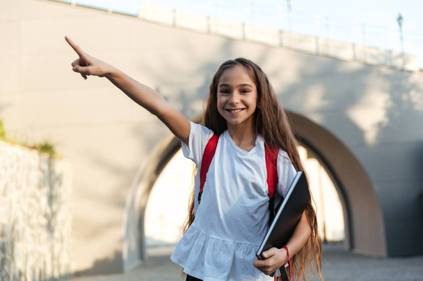Smiling brunette schoolgirl posing outdoors and pointing away - Foto, afbeelding