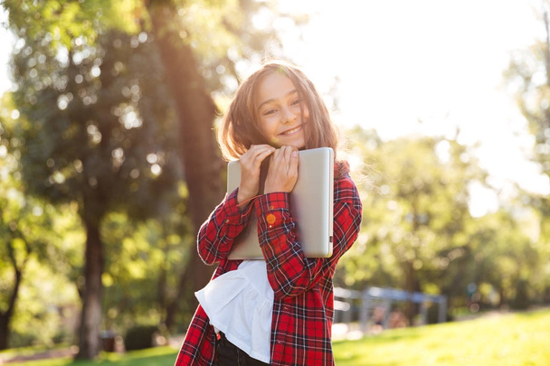 Happy young girl standing in park while hugging her laptop - Zdjęcie, obraz
