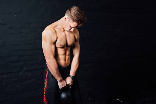 Young muscular man training with kettlebells. man with naked torso on dark background. - Foto, afbeelding