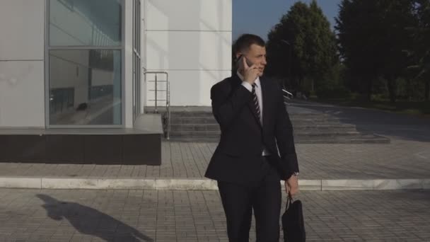 Σοβαρό επιχειρηματία όμορφος σε μαύρο κοστούμι με διπλωμάτης μιλώντας στο smartphone και να σπεύσει σε σύσκεψη ενάντια στο σκηνικό του επιχειρηματικού κέντρου εξωτερική - Πλάνα, βίντεο