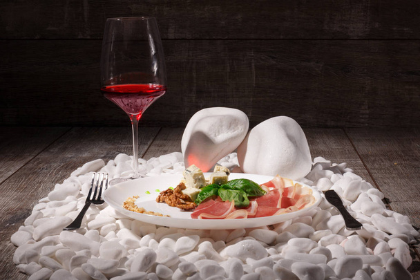 A luxurious composition of a glass of wine and restaurant lunch on a plate on a pile of white rocks. Walnuts, ham and white cheese with fork and knife on a brown wooden background. - Zdjęcie, obraz