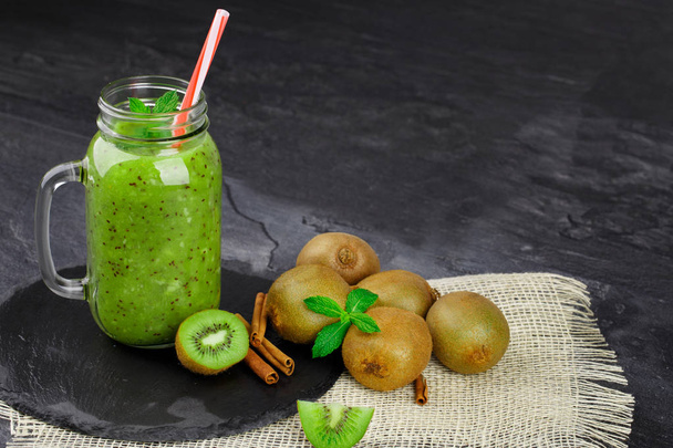 A colorful set of a group of raw kiwis, mason jar full of green milkshake, spicy cinnamon sticks on a white fabric on a stone table background. Juicy ingredients for vegan drinks. Copy space. - Foto, imagen
