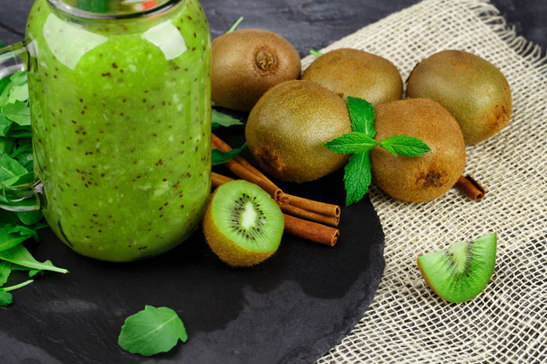 A colorful close-up of a glass jar of kiwi yogurt and healthy ingredients on a fabric on a black background. Decorative mint and aromatic cinnamon next to a pile of kiwi fruits. - Valokuva, kuva