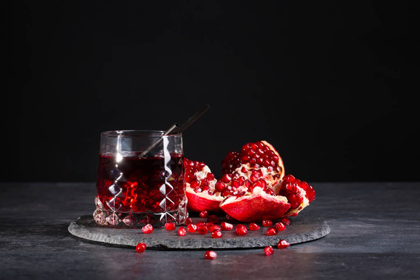 A transparent glass of healthy pomegranate juice and a cut garnet on a round plate on a saturated black background. Organic summer fruity red cocktail with straws and fresh garnet. - Foto, imagen