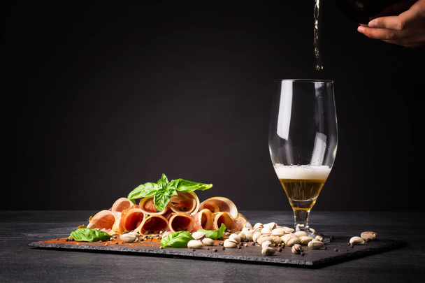 A man pours intoxicating beer into a transparent glass on a saturated black background. An Italian drying balyk in thin slices with green basil near the glass. Prosciutto and pistachios on the table. - Photo, Image